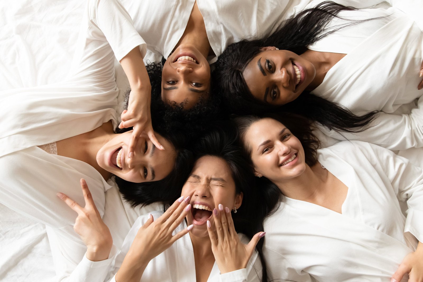 Five happy pretty diverse young girls friends wear robes lying in circle on bed laughing having fun, smiling multi ethnic ladies looking at camera celebrate hen bachelorette party, top view portrait